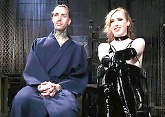 Shiri Allwood sits on a throne in the centre of her dungeon with her captive servant, Ruckus next to her, locked in a cage. This babe wears ebony late