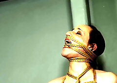 Serf Mila is restrained in unrelenting rope servitude, her body wrapped tightly from head to toe. Her legs, belly, tits, neck, and even her throat are