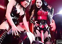 2 dominatrixes in ebony latex corsets and petticoat stand over a stud in a ebony latex straitjacket, thrilling his head and riding him with a thong-on